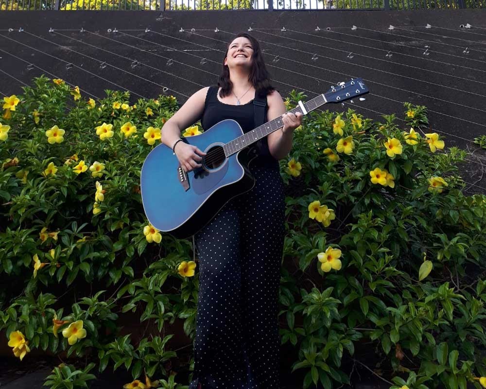 chica morena con un vestido largo negro y una guitarra de color azul y detrás flores de color amarillo